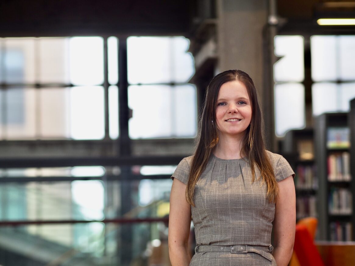 Vrouw in geruit jurkje, lachend, achtergrond met boekenkast en raam.