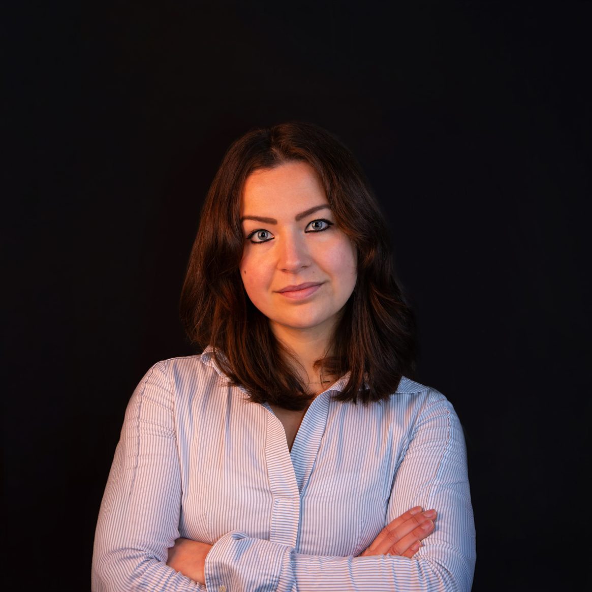 Vrouw met gekruiste armen, gestreepte blouse, donker haar, donkere achtergrond.