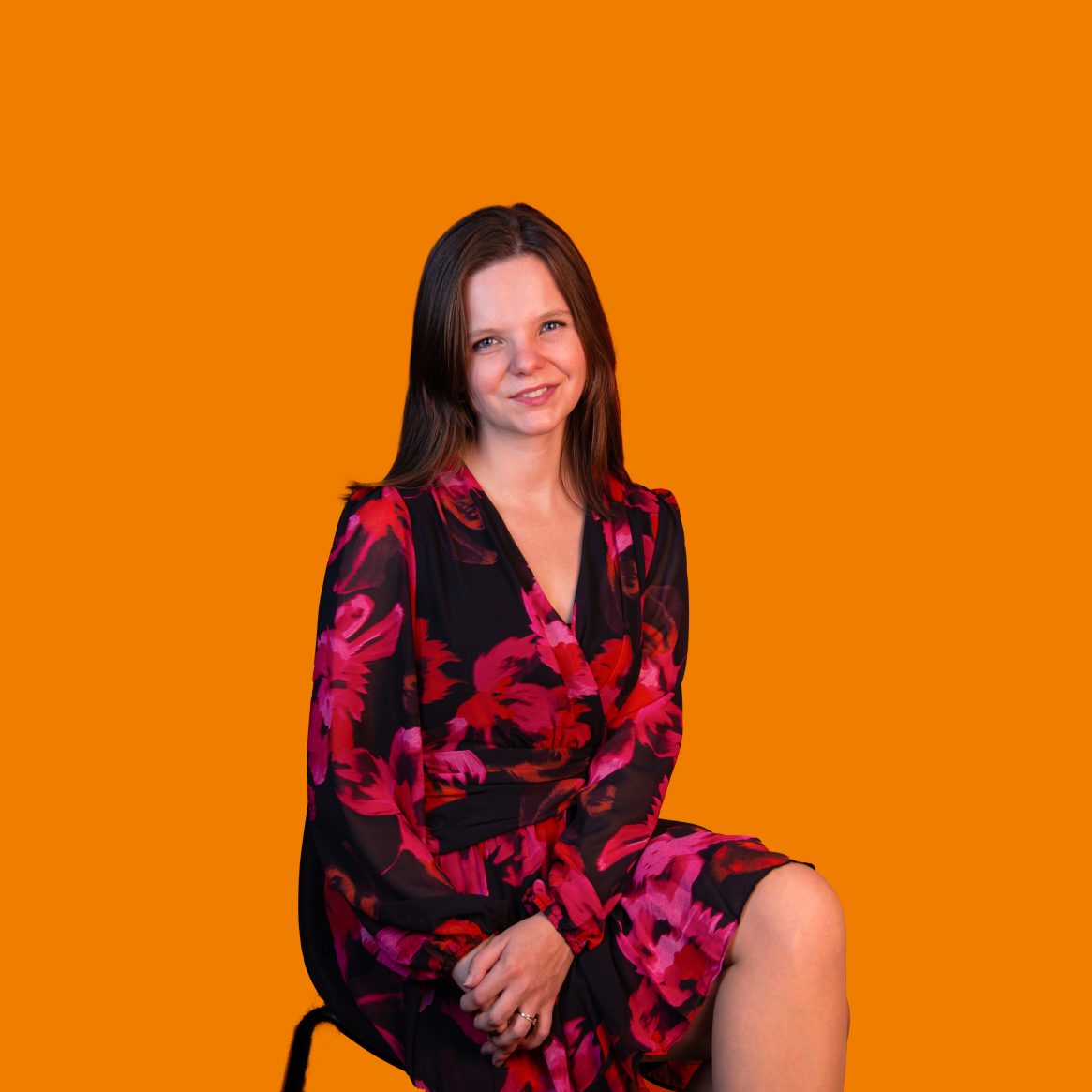 Vrouw in bloemenprintjurk zittend op stoel tegen oranje achtergrond.